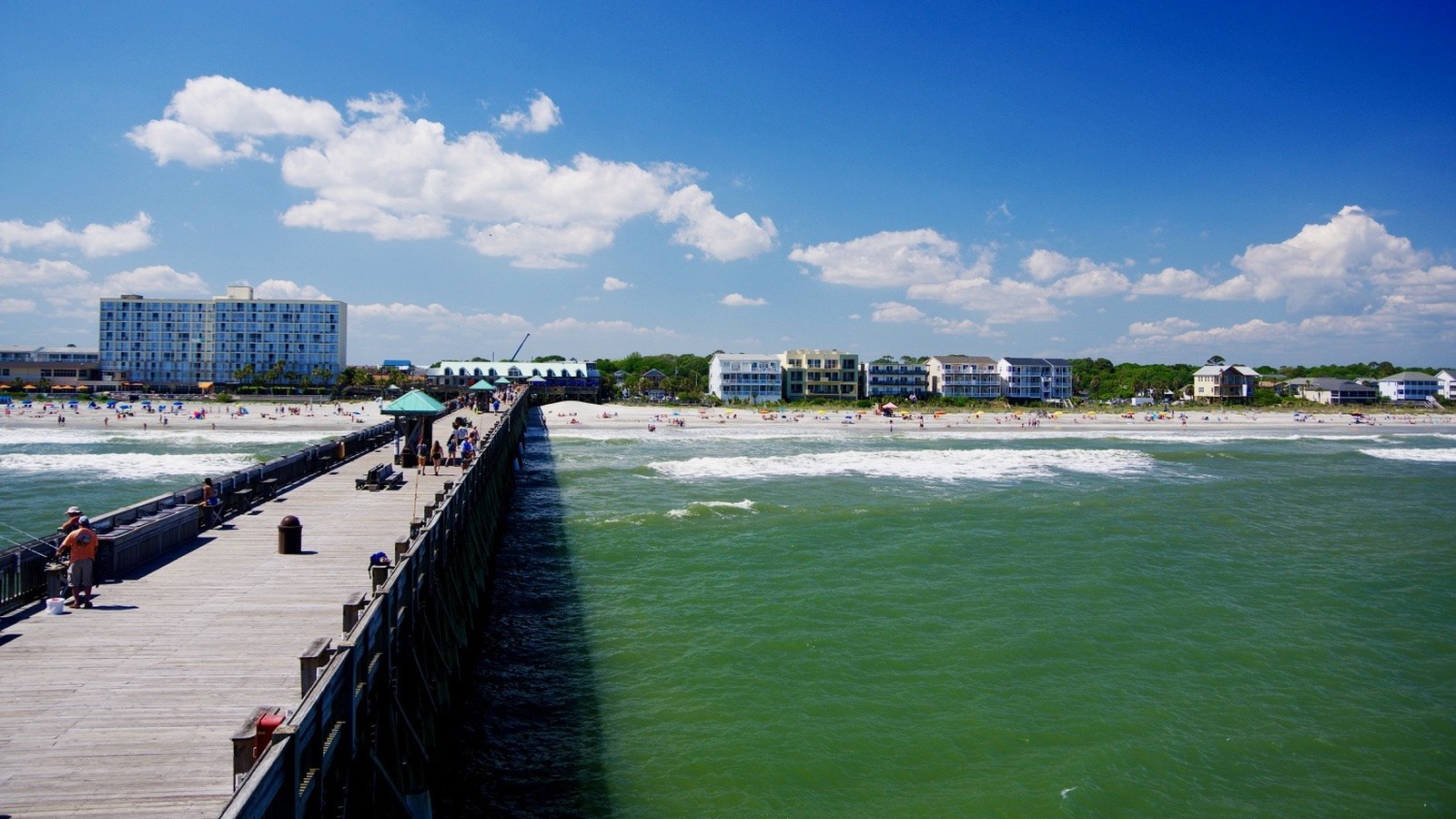 Sunny view down the Folly Beach Pier, with calm green water and the beachfront skyline in the distance