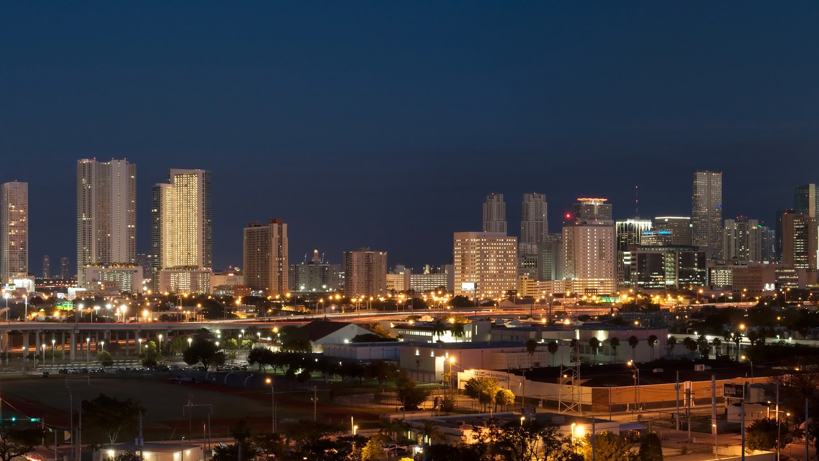 Downtown Miami's high-rise skyline lit up at night, seen across the highway interchange and rail yards to its west