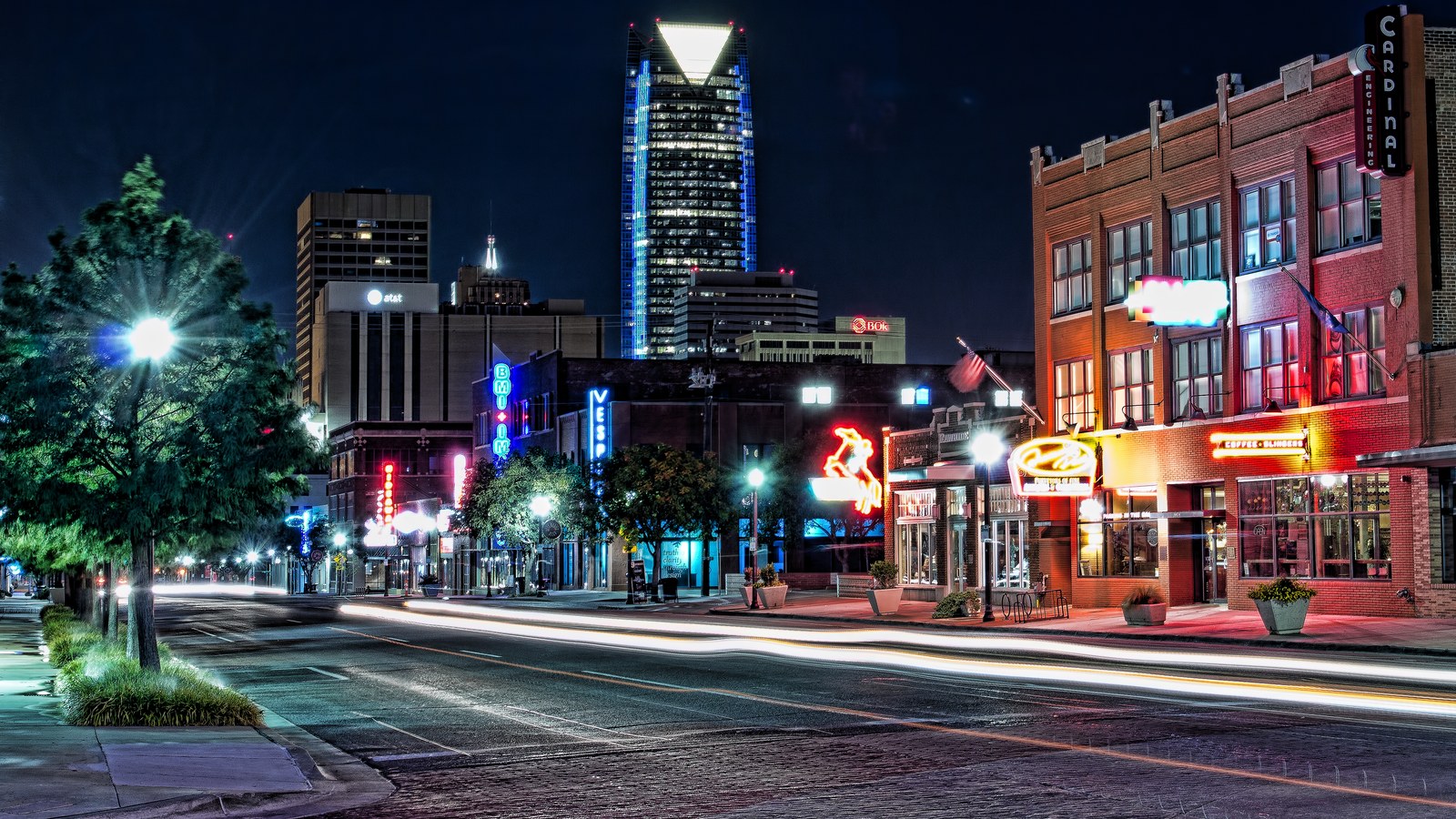 Neon signs lighting up Automobile Alley in Midtown Oklahoma City at night, with the downtown skyline in the background
