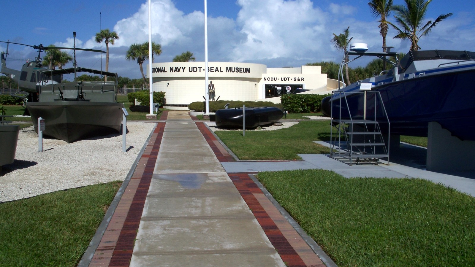 The entrance walkway to the National Navy UDT-SEAL Museum in Fort Pierce, flanked by a helicopter, patrol boats, and other military vessels on outdoor display