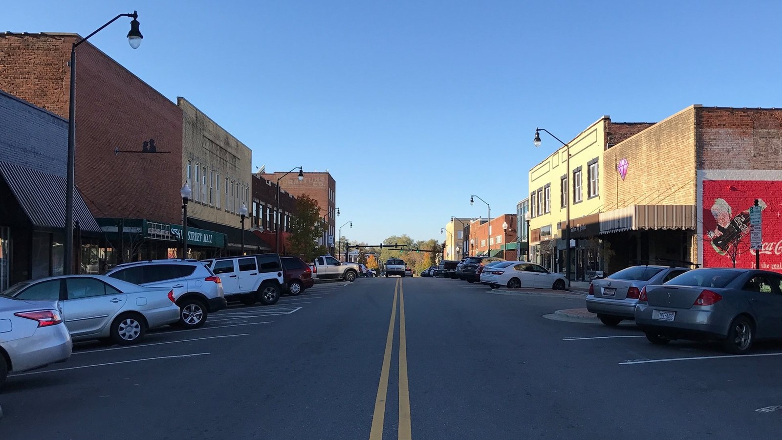 Steele Street in downtown Sanford, with brick storefronts, parked cars, and a Coca-Cola mural on a building at the end of the block