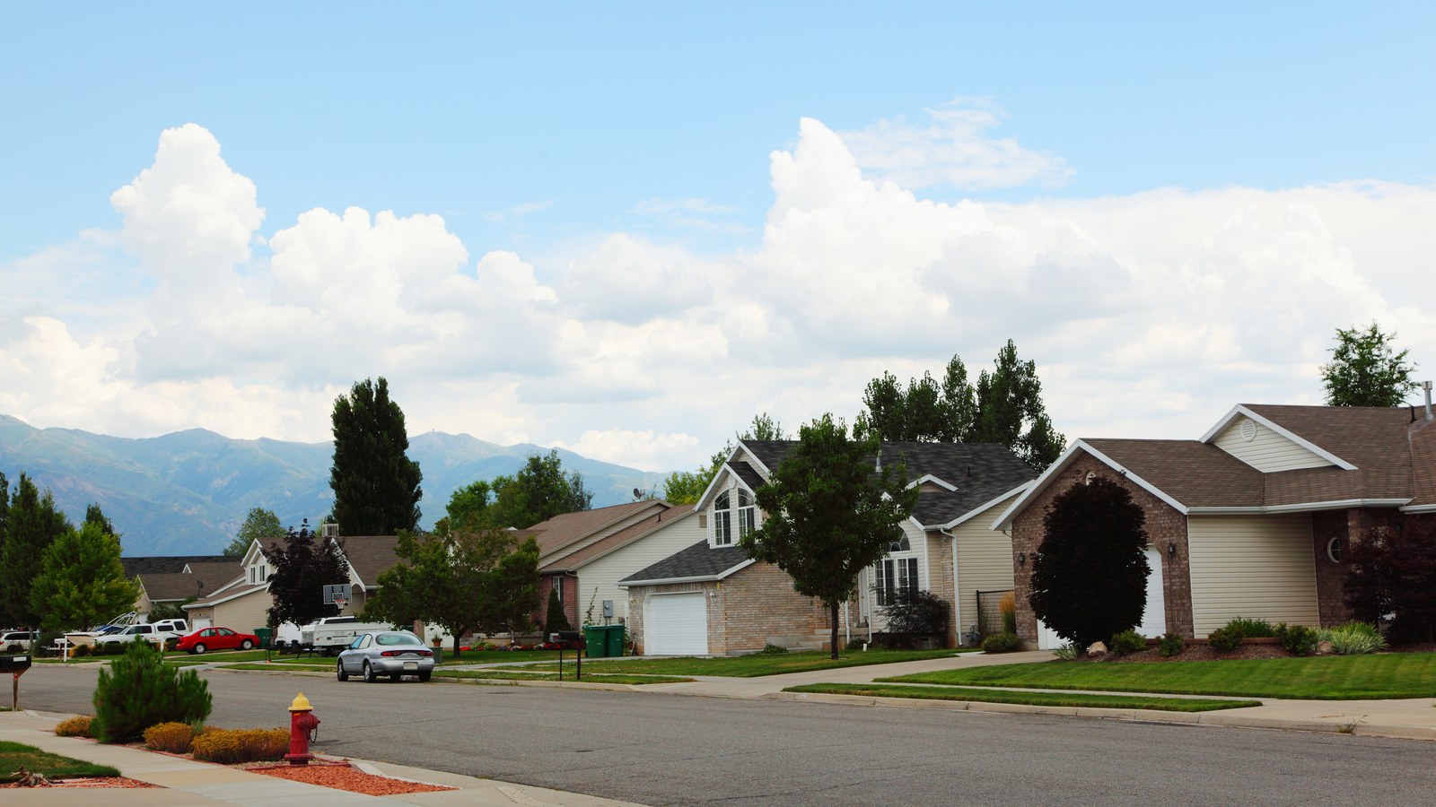 A suburban street of single-family homes in Layton, Utah, with driveways, a fire hydrant, and the Wasatch Range visible in the distance
