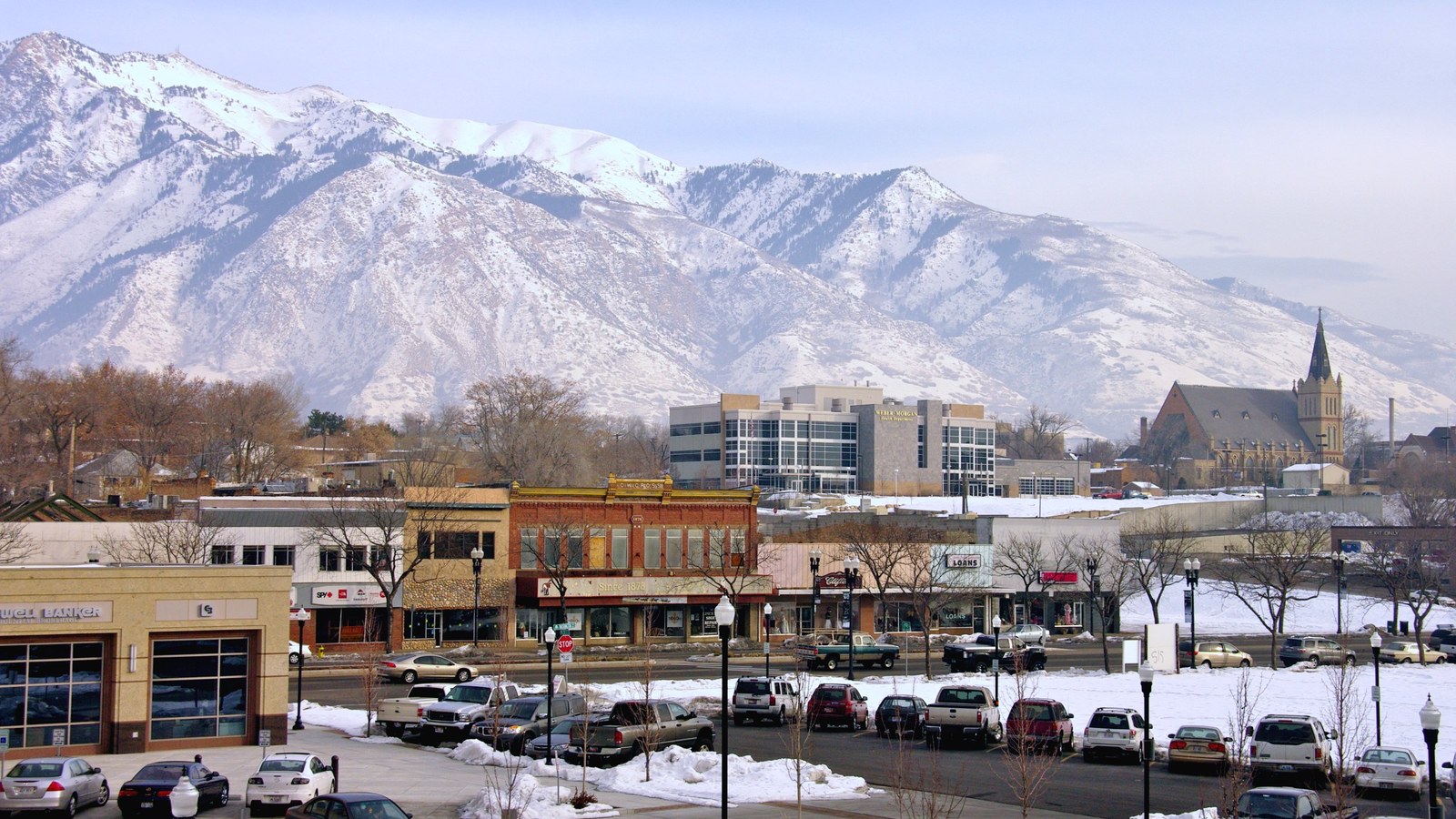 Historic brick storefronts along downtown Ogden, Utah in winter snow, with the Peery's Egyptian Theater district and a church steeple below the snow-covered Wasatch Range
