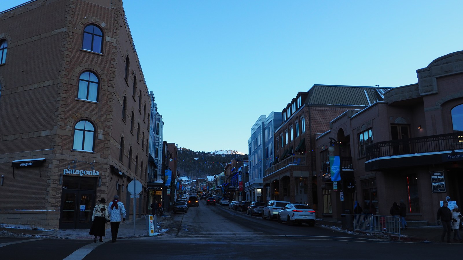Main Street in Park City, Utah at dusk in winter, with historic brick storefronts, string lights, and a snow-covered peak visible at the end of the street